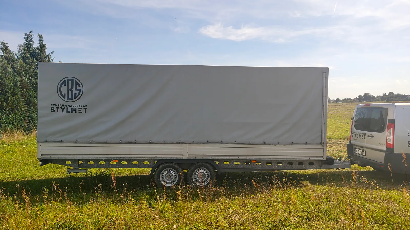 Przyczepa 2700 kg z szarą plandeką w widoku z boku, zaczepiona za samochodem dostawczym